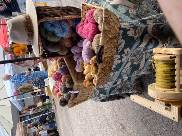 A photo of several baskets of naturally dyed yarn in the Wooly Witch of the West booth at a farmers market.  A brown felt cowgirl hat with a feather band sits on top of the basket and you can see people shopping in the background